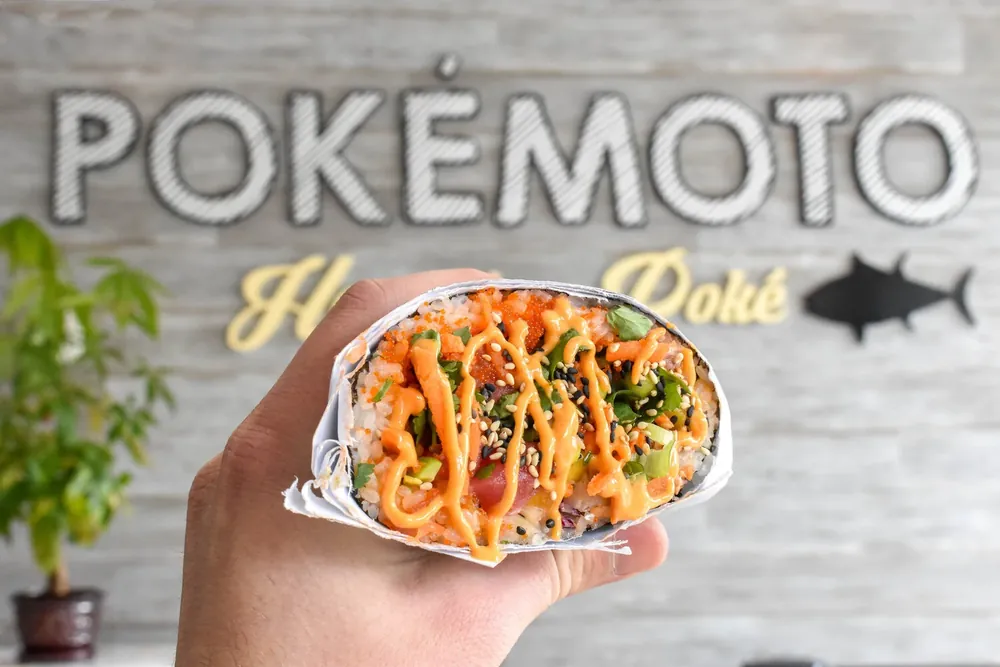 A hand holding a filled poke burrito in front of the Pokemoto sign